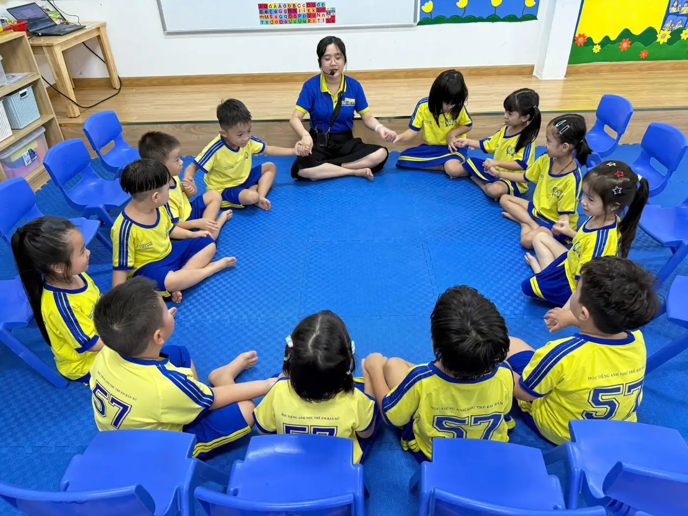 Cô giáo ngồi cùng trẻ lớp Lá thành vòng tròn trên thảm xanh, mọi người nắm tay nhau trong Morning Meeting
