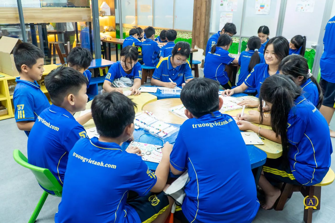 Học sinh tiểu học Trường Việt Anh ngồi quanh bàn lớn trong phòng chế tạo, mỗi nhóm một khay vật liệu và bảng màu